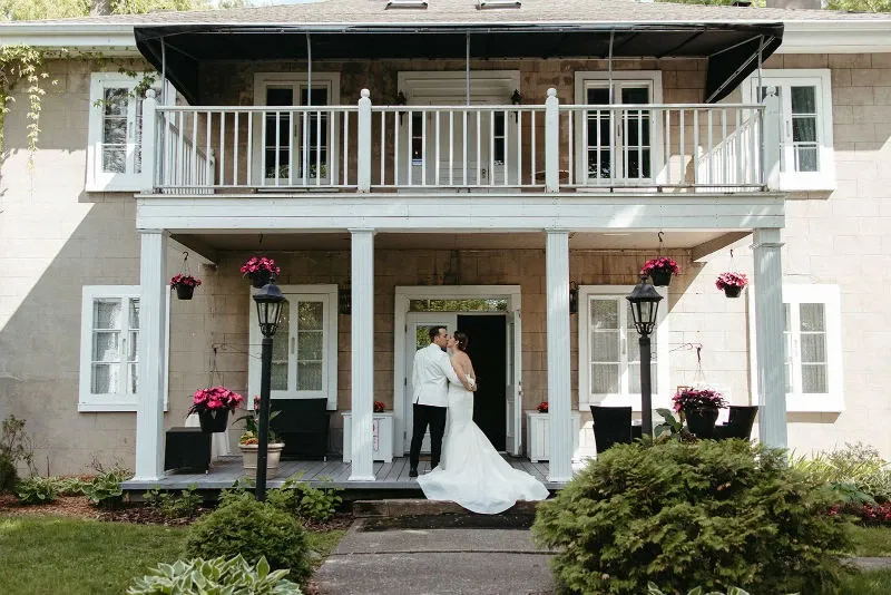 The image shows a couple standing on the porch of the Manoir Grant, a wedding venue listed under the wedding category on Mariage Elegant, Canada. The bride is wearing an elegant white gown, while the groom is in a sophisticated white suit jacket and black trousers. The historic building features a charming two-story design with white-framed windows and a white railing on the balcony. Potted pink flowers adorn the porch, adding a touch of color. Two classic-style black lamps flank the entrance, enhancing the venue's romantic ambiance suitable for a wedding celebration.