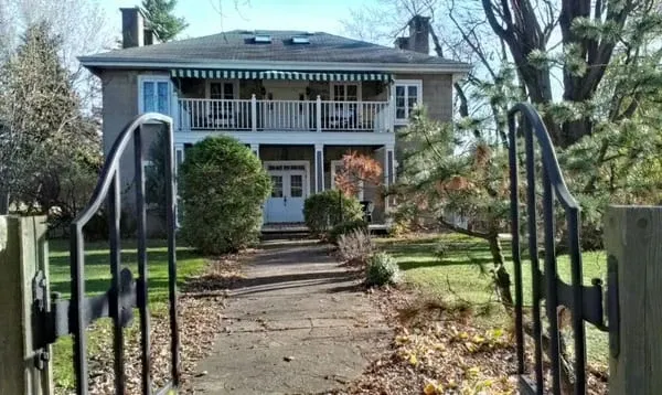The image shows Manoir Grant, a charming wedding venue in Canada, featuring a two-story house with a classic stone facade. The house has a broad balcony on the upper level, adorned with a striped green and white awning. A manicured lawn and garden with bushes and trees surround the pathway leading to the entrance, creating a picturesque and inviting atmosphere. An open wrought-iron gate frames the path, adding to the venue's elegant appeal, ideal for wedding ceremonies and receptions.
