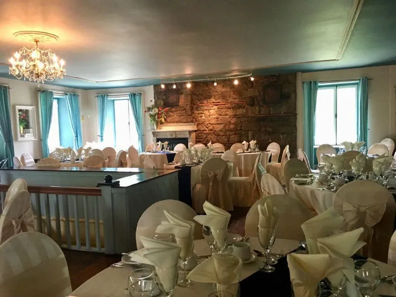 The image shows an elegantly decorated indoor wedding venue at Manoir Grant, a featured vendor in the Wedding Venues category for Mariage Elegant, Canada. The room is adorned with round tables covered in white linens, each set with neatly arranged glassware and silverware. Chairs are draped with white covers and tied with light blue sashes, complementing the soft blue curtains framing the windows. A grand chandelier hangs from the ceiling, casting a warm glow over the space. At the far end, a rustic stone fireplace adds a charming, cozy touch to the wedding setting.