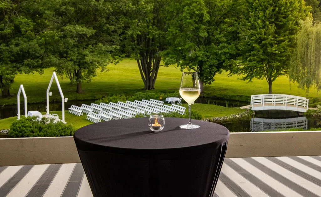 This image features The Alto & Soprano, a wedding venue in Canada, showcasing an elegant outdoor ceremony setup. Rows of white chairs are arranged on a lush green lawn, facing a modern white arch for the wedding ceremony. In the foreground, a small black table is adorned with a glass of white wine and a tealight candle, adding a touch of sophistication. A picturesque white bridge spans across a serene pond amidst a backdrop of vibrant greenery. This venue provides a romantic and picturesque setting for weddings.