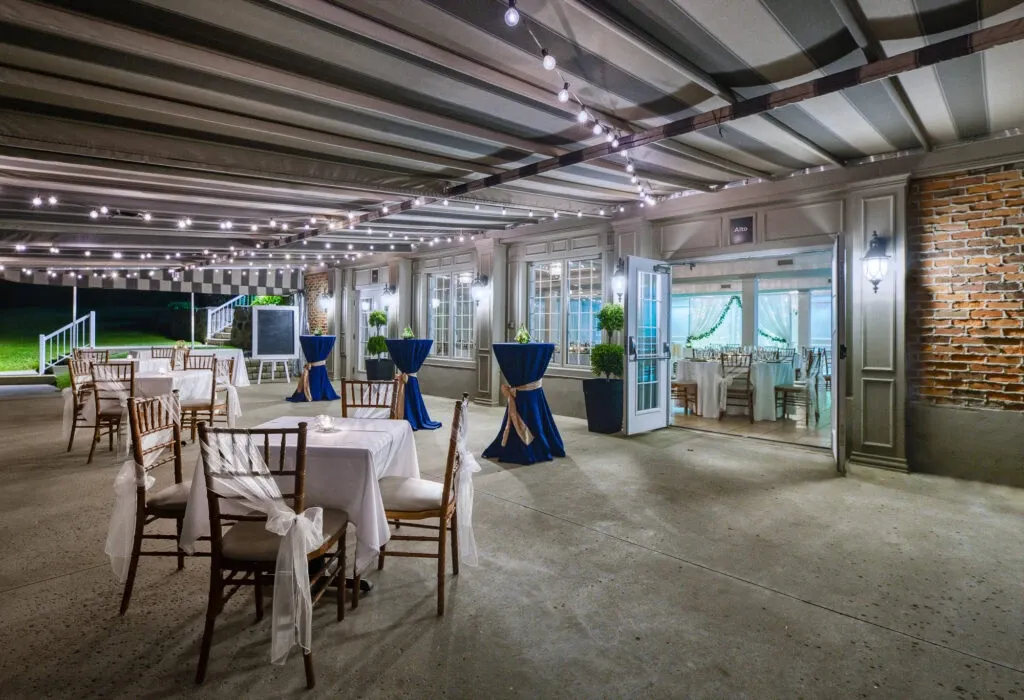 The image showcases The Alto & Soprano, a wedding venue in Canada, listed under Mariage Elegant's vendor offerings. The venue features a spacious outdoor area elegantly set up for a wedding event. Rows of white-clothed tables surrounded by wooden chairs with sheer white covers are strategically placed under a charming pergola adorned with strings of twinkling lights. Tall, blue-draped cocktail tables intersperse the space, adding a touch of sophistication. A brick and paneled backdrop enhances the area, while lush greenery peeks from behind glass doors, offering a glimpse of a serene indoor space. The ambiance is inviting and romantic, ideal for a memorable celebration.