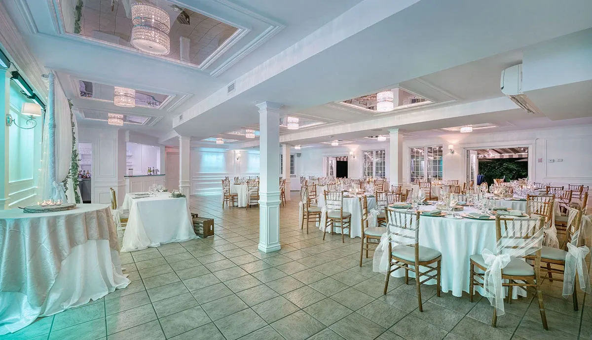 The image showcases The Alto & Soprano, a distinguished venue listed under wedding venues on Mariage Elegant, Canada. The spacious room features an elegant setup with neatly arranged round tables draped in white linens and adorned with delicate centerpieces. Chiavari chairs are positioned around each table, adding a touch of sophistication. Soft, ambient lighting highlights the classic architectural details, including ornate ceilings with embedded chandeliers. A designated area for a dance floor or additional seating is visible, suggesting versatility for wedding receptions. The overall ambiance reflects a serene and romantic environment, ideal for wedding celebrations.