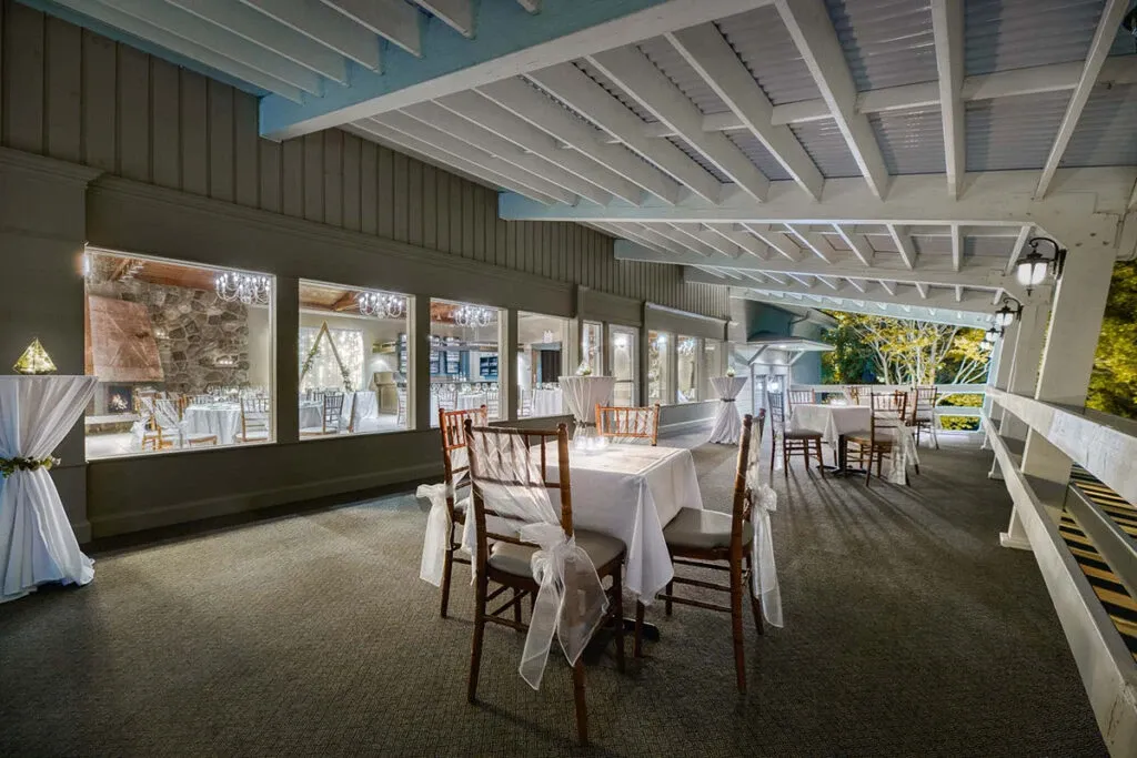 This image showcases The Alto & Soprano, a distinguished wedding venue listed under Wedding Venues at Mariage Elegant, Canada. The setting features an elegantly decorated patio with wooden chairs wrapped in sheer white fabric, enhancing the romantic ambiance. Large windows line the right side, offering a view of the indoor area adorned with chandeliers and stone wall elements, creating a blend of rustic and refined charm. The outdoor space is set up with tables covered in white linen, perfect for a serene and intimate wedding reception.