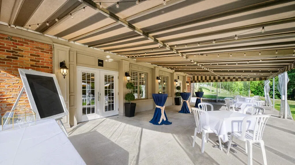 The image showcases a beautifully arranged outdoor wedding venue by The Alto & Soprano, featured on Mariage Elegant's Canada vendor listing. The setting includes a covered patio with a striped awning, offering a shady space for guests. White tables and chairs are neatly arranged, accompanied by elegant blue tablecloths on standing cocktail tables, creating a sophisticated and inviting atmosphere. The backdrop features a charming brick and cream facade with large windows, adorned with small potted plants, adding a touch of greenery. The venue exudes a picturesque and tranquil ambiance, perfect for a wedding celebration.