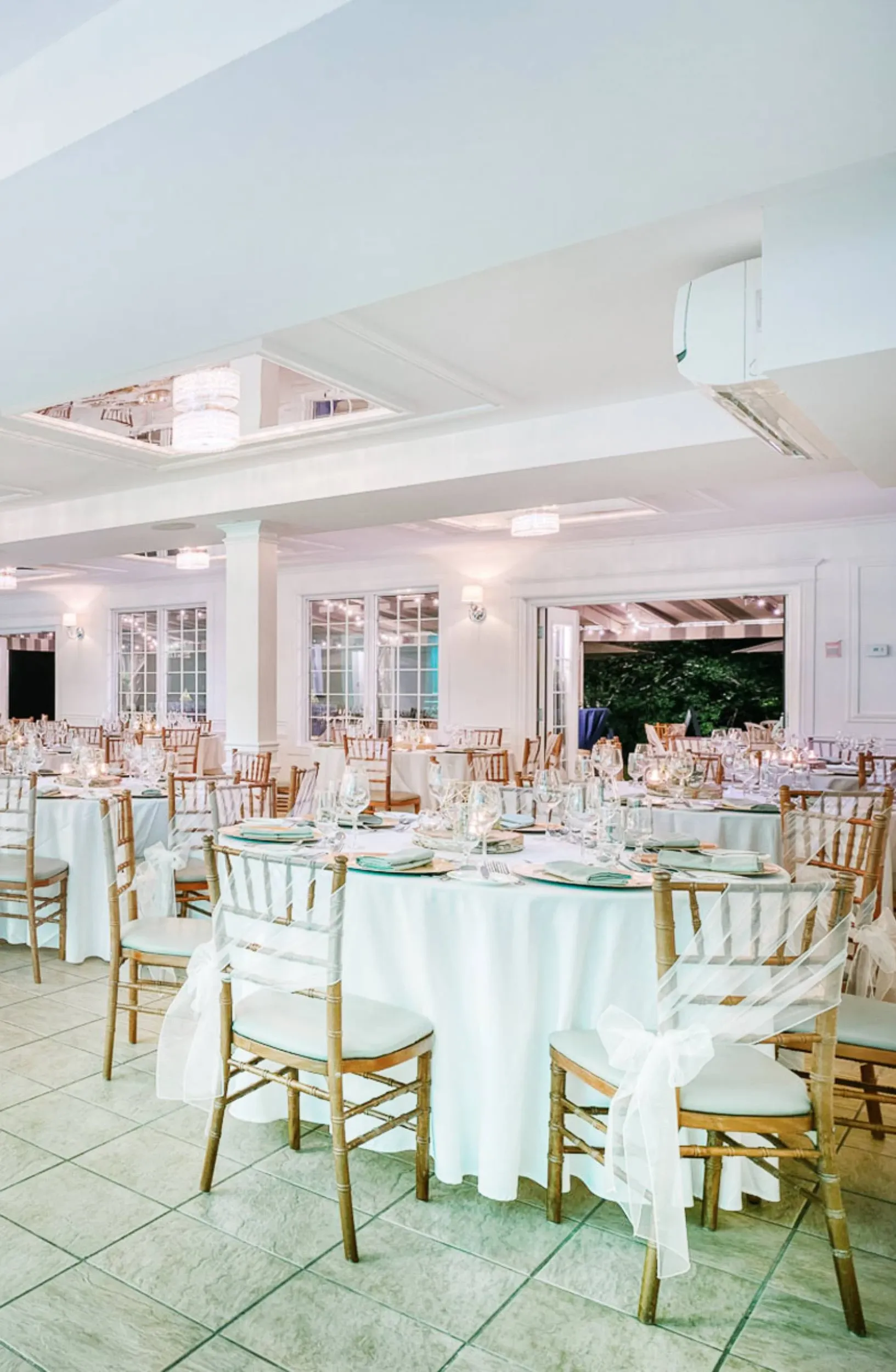 The image showcases a beautifully arranged interior of a wedding venue by The Alto & Soprano, categorized under Wedding Venues, available on Mariage Elegant, Canada. The room features elegantly set round tables draped in white linens, complemented by wooden chairs adorned with sheer white sashes. Each table is meticulously prepared with fine china, sparkling glassware, and neatly arranged silverware, creating a sophisticated ambiance. Soft lighting and large windows allow natural light to fill the space, enhancing the romantic atmosphere ideal for a wedding celebration.