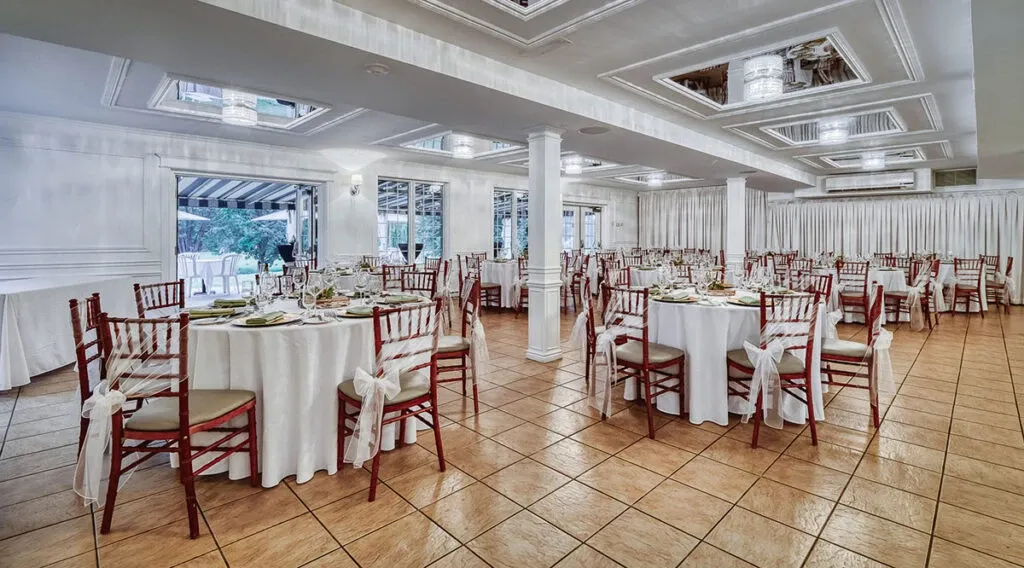 A beautifully decorated wedding venue, The Alto & Soprano, is set up for a reception. The spacious room features elegantly arranged round tables with white tablecloths and delicate place settings, showcasing the venue's attention to detail. Wooden chairs with white sashes surround each table, adding a touch of sophistication. The ceiling has modern light fixtures that illuminate the space with a soft glow. Large windows on one side let in natural light, offering a glimpse of greenery outside. This image highlights the serene and elegant atmosphere perfect for any wedding celebration at this Canadian venue, listed under the wedding venue category on Mariage Elegant.