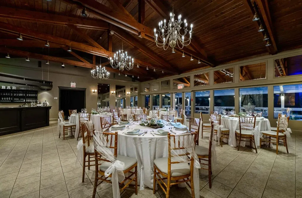 The image showcases the interior of 'The Alto & Soprano,' a wedding venue in Canada, listed on Mariage Elegant. The spacious room features elegantly set round tables adorned with white tablecloths and decorative centerpieces, complemented by wooden Chiavari chairs draped with white fabric sashes. The ceiling boasts exposed wooden beams and several hanging chandeliers, creating a warm and inviting atmosphere. Large windows line one side of the room, offering ample natural light and a view of the outdoor landscape. The venue exudes a charming and sophisticated ambiance ideal for wedding celebrations.