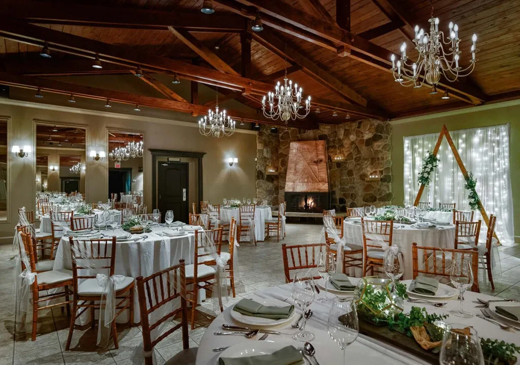 The image showcases The Alto & Soprano, a wedding venue under the category of Wedding Venues, featured on Mariage Elegant, Canada. The venue's spacious interior is elegantly arranged with round tables draped in white linens and accompanied by wooden chairs. Each table is adorned with neatly arranged cutlery, wine glasses, and fresh green foliage as centerpieces, adding a touch of nature. The room's rustic charm is enhanced by a prominent stone fireplace centrally positioned, framed by a warm glow. Overhead, chandeliers and exposed wooden beams contribute to the inviting ambiance. A backdrop of soft, white string lights and greenery adds a romantic touch, suitable for a memorable wedding celebration.