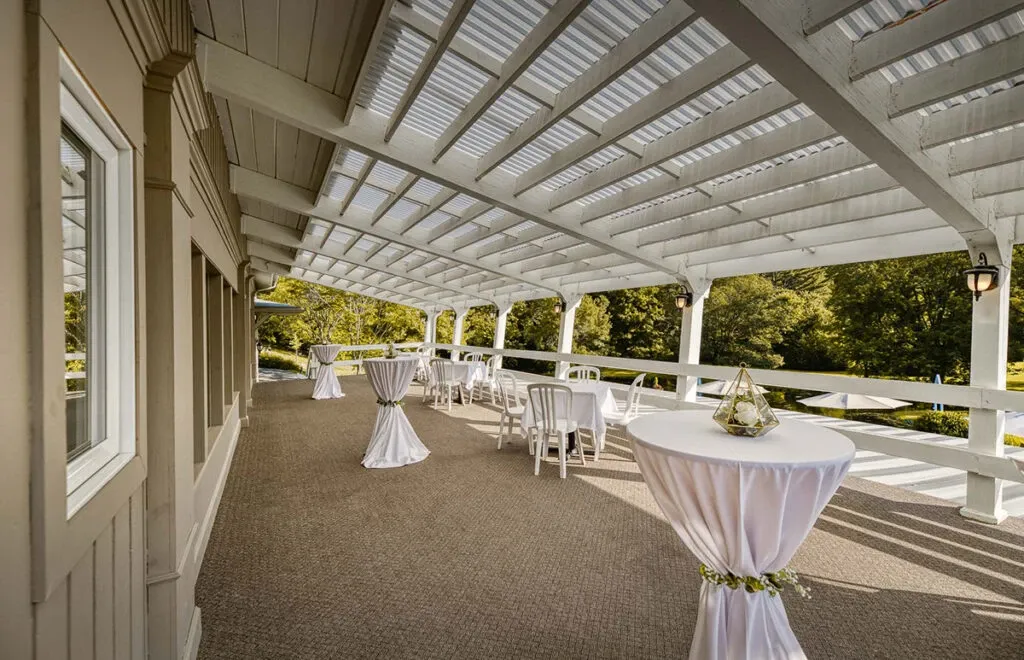 The image showcases an elegant outdoor area belonging to The Alto & Soprano, categorized under wedding venues on Mariage Elegant, Canada. The venue features a spacious, covered patio with white pergola roofing and a beige exterior facade, providing a serene and stylish setting suitable for wedding events. Several cocktail tables are adorned with white linens, and one table is tastefully decorated with a glass terrarium centerpiece. White chairs are arranged neatly beside some tables, suggesting readiness for guests to enjoy. The surrounding lush greenery adds a natural and inviting backdrop, ideal for hosting memorable weddings in a picturesque environment.