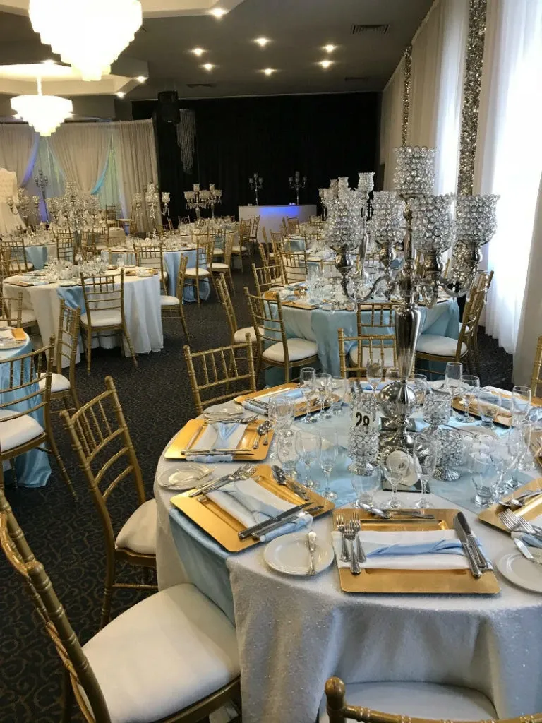 The image shows an elegantly decorated wedding venue at the Renaissance Centre, listed under the wedding category. It features round tables draped in white tablecloths with light blue accents, adorned with tall, intricate crystal candelabras as centerpieces. The tables are set with fine tableware, including plates, silverware, and neatly folded napkins. Gold Chiavari chairs surround the tables, enhancing the luxurious atmosphere. The venue's lighting is soft and ambient, complemented by chandeliers hanging from the ceiling. The draped fabric and stylish decor contribute to a sophisticated and romantic setting for a wedding celebration.