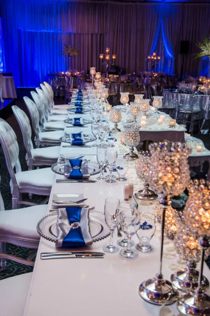 The image showcases an elegant wedding reception setting at Renaissance Centre, categorized under Wedding Venues for Mariage Elegant in Canada. The venue features a long, beautifully decorated banquet table adorned with gleaming crystal candle holders and shimmering votive candles, creating a romantic atmosphere. Each place setting is meticulously arranged with a silver beaded charger plate, fine glassware, and a navy blue napkin, elegantly folded with a sparkling embellishment. Plush white chairs line either side of the table, further contributing to the sophisticated ambiance. Ambient blue lighting bathes the room, enhancing the luxurious and festive environment ideal for a memorable wedding celebration.