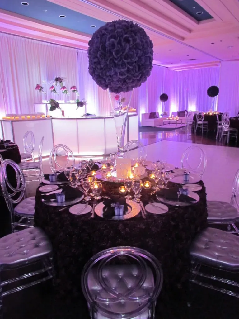 The image showcases an elegantly set wedding reception table at the Renaissance Centre, a premier wedding venue listed on Mariage Elegant, Canada. The table features a luxurious, dark lace tablecloth complemented by transparent ghost chairs, creating a modern and sophisticated ambiance. A striking tall centerpiece with a lush, dark floral arrangement dominates the table, surrounded by sparkling glassware and glowing candlelight, contributing to the romantic atmosphere. In the background, there is soft purple ambient lighting, enhancing the overall elegant and inviting setting ideal for a wedding celebration.