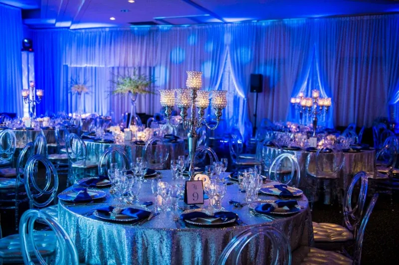 The image showcases an elegant wedding reception setup at the Renaissance Centre, categorized under wedding venues for Mariage Elegant, Canada. The venue is beautifully decorated with round tables covered in shimmering silver tablecloths, each adorned with sparkling crystal candelabras as centerpieces. The tables are set with fine glassware, white and blue floral arrangements, and neatly folded blue napkins, creating a sophisticated atmosphere. The room is illuminated with soft blue lighting, adding a romantic ambiance, while white drapery lines the walls, enhancing the venue's luxurious feel.