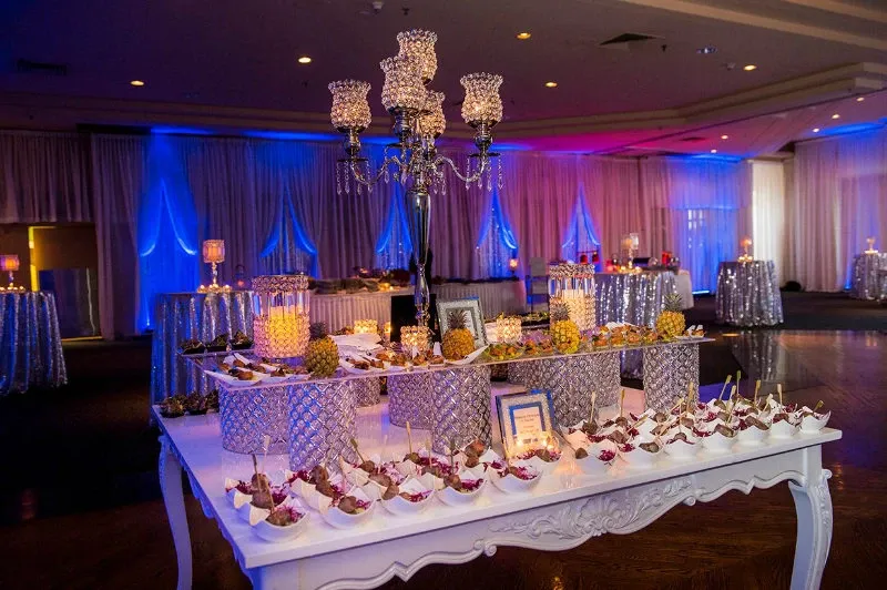 The image shows an elegantly set up reception area at the Renaissance Centre, a prominent wedding venue in Canada, under the wedding planning category. The room features dim lighting with blue and purple hues creating a sophisticated ambiance. At the center is a white ornate table adorned with intricate metal candle holders and an assortment of appetizers artfully displayed on small plates. In the background, the tables are draped with shimmering silver tablecloths and decorated with tall candle holders, adding to the luxurious setting ideal for a wedding celebration.