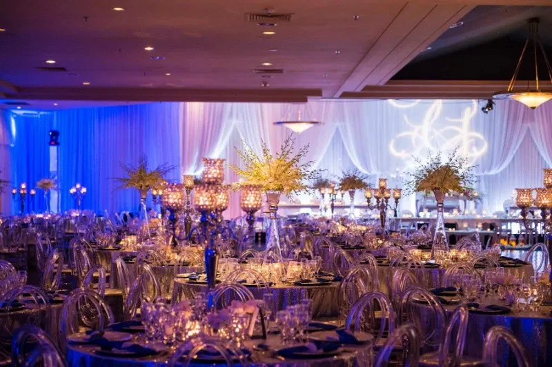 The image showcases a beautifully decorated wedding venue at the Renaissance Centre, a featured wedding venue in the Mariage Elegant vendor listing in Canada. The room is elegantly lit with a soft blue ambiance, highlighting lavish table settings adorned with crystal glassware and tall floral centerpieces. The backdrop features sheer drapes with subtle lighting, creating a romantic and sophisticated atmosphere perfect for a wedding reception. The venue's elegant design reflects a luxurious setting ideal for hosting memorable wedding events.