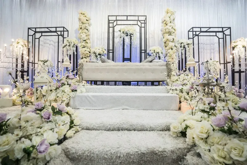 The image shows an elegant wedding venue setup at the Renaissance Centre, categorized as a wedding venue on Mariage Elegant, Canada. The scene features a luxurious stage decorated with abundant white and pastel flowers, including roses and hydrangeas, beautifully arranged around ornate metal frames. In the center, there is a sophisticated white cushioned sofa, surrounded by tall floral arrangements and soft fabric draping the backdrop, creating a romantic atmosphere. The soft lighting enhances the serene and enchanting ambiance, perfect for a wedding ceremony or reception.