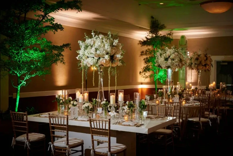 This image features a beautifully decorated wedding venue at the Renaissance Centre, categorized under Wedding Venues on Mariage Elegant, Canada. The setting showcases an elegantly arranged reception area with a long dining table adorned with tall floral centerpieces made of white and cream flowers. The table is set with fine china, glassware, and numerous candles that add a romantic glow to the ambiance. Chiavari chairs with white cushions surround the table. Lush greenery, including small trees with soft, green lighting, enhances the sophisticated and enchanting atmosphere of the venue, perfect for a memorable wedding celebration.
