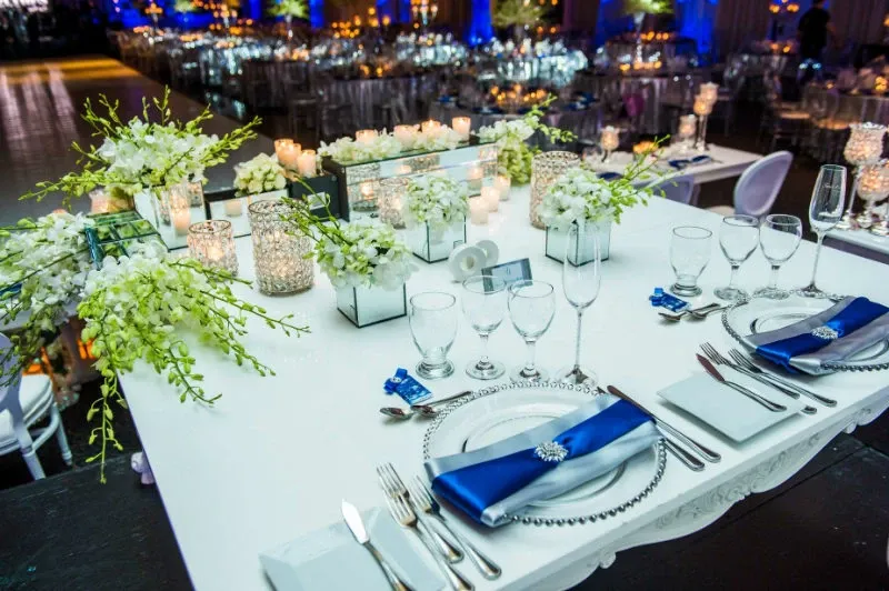 The image showcases a beautifully decorated wedding reception table at Renaissance Centre, categorized under Wedding Venues on Mariage Elegant, Canada. The table features elegant white floral arrangements and a variety of candles adding a romantic ambiance. Each place setting includes a crystal-clear plate, silver cutlery, and a neatly folded navy blue napkin adorned with a decorative brooch. In the background, more tables are similarly arranged, with gentle blue lighting enhancing the venue's sophisticated atmosphere, making it an ideal setting for elegant wedding celebrations.