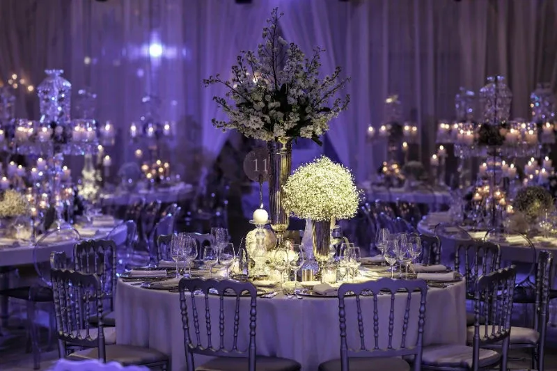 The image showcases an elegantly decorated wedding venue at the Renaissance Centre, categorized under Wedding Venues, for Mariage Elegant in Canada. The setting features round tables adorned with white tablecloths and surrounded by stylish silver chairs. Each table is beautifully arranged with intricate floral centerpieces that include cascading white flowers and greenery, creating a romantic ambiance. Dimly lit, the venue is illuminated by numerous candles and soft lighting, enhancing the intimate and sophisticated atmosphere ideal for a wedding celebration. The draped white curtains in the background add a touch of grace and luxury to the scene.