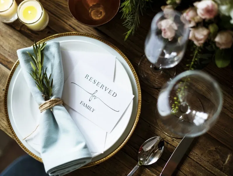A beautifully set wedding table at the Renaissance Centre, showcasing its elegance as a wedding venue. The table features a white plate with a gold rim, topped with a light blue napkin and a sprig of greenery, neatly tied with twine. A card labeled 'Reserved for Family' is elegantly placed on the plate. Surrounding the setting, delicate glassware and polished cutlery are arranged meticulously. Soft candlelight and a floral arrangement in the background contribute to the sophisticated ambiance, perfect for any wedding celebration.
