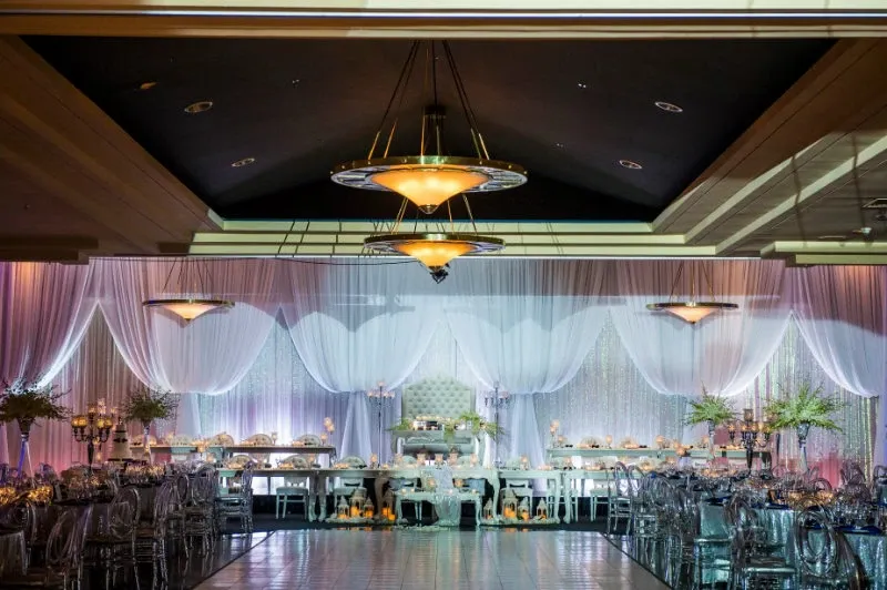 The image showcases an elegantly decorated wedding venue at the Renaissance Centre, a featured wedding venue in the Mariage Elegant, Canada listings. The spacious hall is adorned with soft, flowing white drapes that create a luxurious ambiance. Overhead, warm chandeliers cast a gentle glow over the venue. A long, beautifully set banquet table is positioned prominently, draped with a crisp white tablecloth and decorated with elegant floral arrangements and candleholders, adding a romantic touch. The overall setting exudes sophistication, making it an ideal location for a memorable wedding celebration.