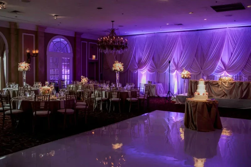 The image showcases an elegant wedding venue at the Renaissance Centre, categorized under wedding venues on the Mariage Elegant, Canada site. The room is beautifully decorated with soft purple lighting that casts a romantic glow over the draped fabric walls. The tables are set with exquisite floral centerpieces, featuring white roses and lush greenery. A polished white dance floor takes center stage, while a charming three-tier wedding cake is displayed on a draped table. The ambiance is further enhanced by a grand chandelier, adding a touch of sophistication to the venue.