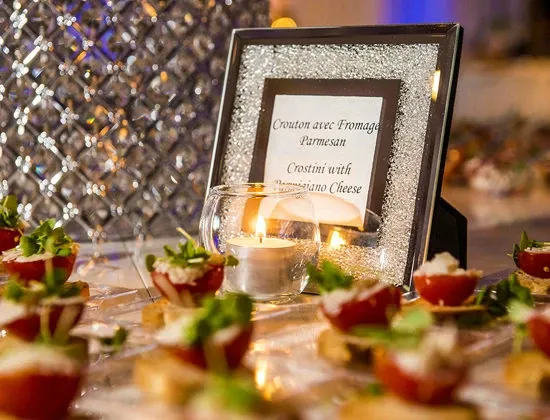 The image shows an elegant table setting at the Renaissance Centre, a wedding venue listed on Mariage Elegant, Canada. In the foreground, there are beautifully arranged appetizers, including tomato halves topped with a garnish and small pieces of crostini with Parmesan cheese. A small glass-encased candle adds a warm ambiance to the scene. A framed sign with the text 'Crostini with Parmesan Cheese' is prominently displayed, suggesting the catering style offered by the venue. The background features a tastefully decorated wall with a textured pattern, enhancing the luxurious atmosphere suitable for weddings.