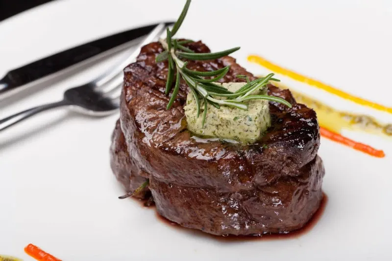The image showcases a beautifully presented gourmet steak dish from the Renaissance Centre, categorized under wedding venues on Mariage Elegant, Canada. The steak is perfectly cooked and topped with a dollop of herbed butter, garnished with a sprig of fresh rosemary. The plate features artistic drizzles of yellow and red sauces, adding a touch of elegance that complements the sophisticated dining experience expected at this wedding venue. A shiny fork and knife are placed neatly beside the dish, completing the elegant table setting.