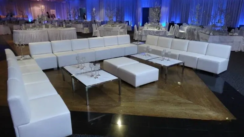 The image depicts a modern wedding venue setup at the Renaissance Centre in Canada under the category of Wedding Venues. The scene features a spacious indoor area with sleek, white leather sectional sofas arranged around low, square tables. Each table is elegantly adorned with clear champagne flutes. The floor has a stylish mix of dark and light tiles, enhancing the contemporary atmosphere. In the background, several dining tables covered in white tablecloths are gracefully set up. The room is decorated with soft blue uplighting and delicate tree branch centerpieces, creating a sophisticated and romantic ambiance perfect for a wedding celebration.