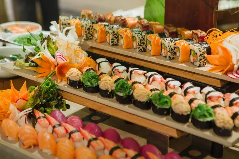 An elegant display of assorted sushi is artfully arranged on tiered wooden platforms, featuring a variety of colorful rolls including salmon, tuna, and nigiri, accented with vibrant garnishes like sliced vegetables and carved carrots. This visually appealing food spread is part of the catering services offered by Renaissance Centre, a top choice for wedding venues in Canada. The presentation highlights the venue's commitment to providing exceptional dining experiences for weddings and special events.