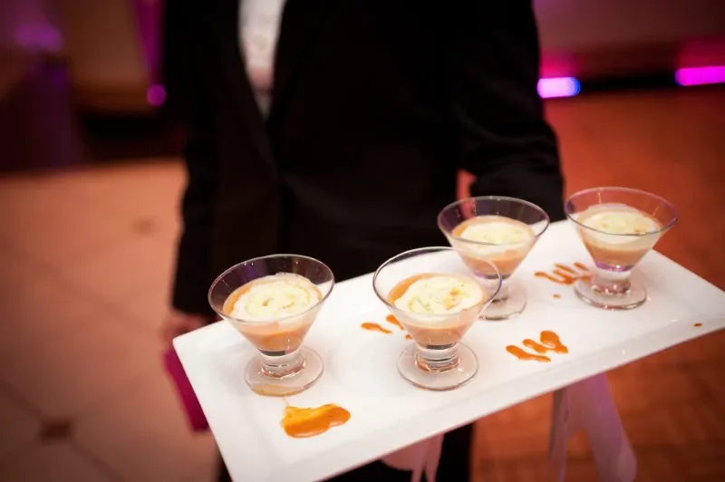 A server dressed in a formal black suit holds a white tray featuring four elegantly presented dessert parfaits in martini glasses, each topped with a creamy layer and a drizzle of caramel sauce. The setting, with ambient pink lighting and a polished wooden floor, suggests a sophisticated atmosphere, suitable for a wedding reception. This image highlights the catering offerings at the Renaissance Centre, a renowned wedding venue in Canada.