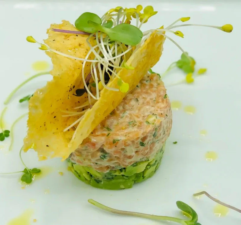 A gourmet dish from Renaissance Centre, a featured wedding venue on Mariage Elegant, Canada, showcasing a beautifully presented stack of finely diced seafood tartare atop a layer of avocado. The dish is artistically garnished with vibrant microgreens and topped with a delicate, crisp parmesan tuile. This elegant culinary presentation reflects the quality catering services available for wedding events at this venue.