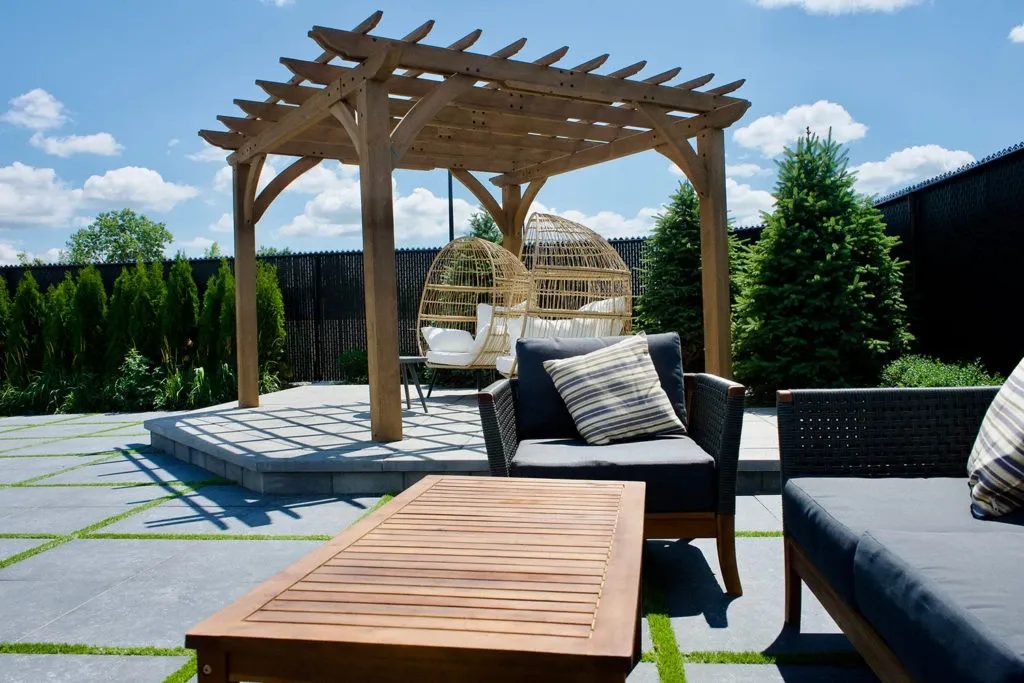 This image showcases the outdoor area of Plaza Volare, categorized as a wedding venue, perfect for elegant ceremonies. The setting features a charming wooden pergola on a raised stone platform, providing an ideal focal point for wedding vows. Underneath the pergola are two cozy, egg-shaped wicker chairs, creating a romantic nook for couples. The surrounding landscape includes lush green trees and neatly trimmed hedges, adding to the serene atmosphere. In the foreground, modern patio furniture with cushioned seating and a wooden coffee table offer a comfortable space for guests to relax amidst the picturesque setting.