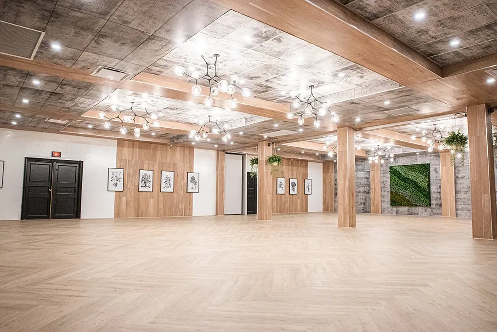 This image features the elegant interior of Plaza Volare, showcasing it as a premier wedding venue. The spacious room is adorned with modern chandeliers hanging from the ceiling, casting a warm glow over the light-colored, polished hardwood floor. Wooden beams and paneling add a touch of rustic charm, complemented by framed artwork lining the walls. A striking green plant wall serves as a focal point, enhancing the sophisticated and natural ambiance. This setting highlights Plaza Volare's potential to host beautifully memorable wedding celebrations.