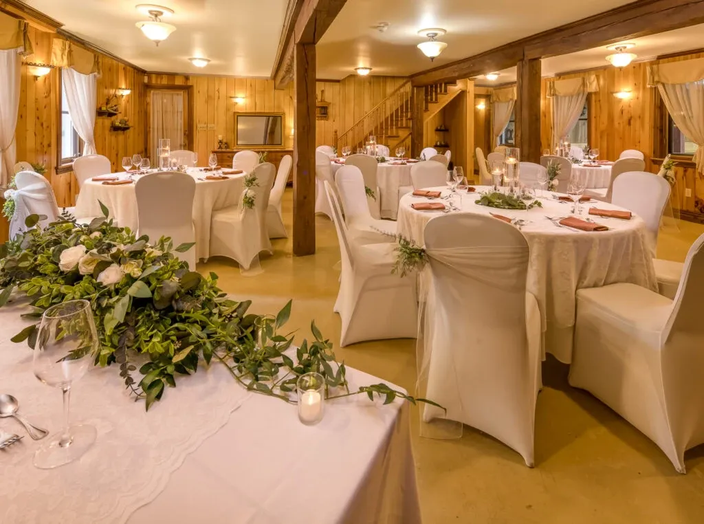 The image shows the interior of L’Érablière D’autrefois Hall, a wedding venue featured on Mariage Elegant, Canada. The rustic hall is adorned with wooden walls and beams, creating a warm and inviting atmosphere. Round tables are elegantly set for a wedding reception, covered with white tablecloths and surrounded by white chair covers. Each table has a simple floral centerpiece made of greenery and white roses, enhancing the natural, elegant decor. The soft lighting and curtains add a cozy and intimate feel to the space, making it a perfect choice for couples looking for a charming wedding venue.