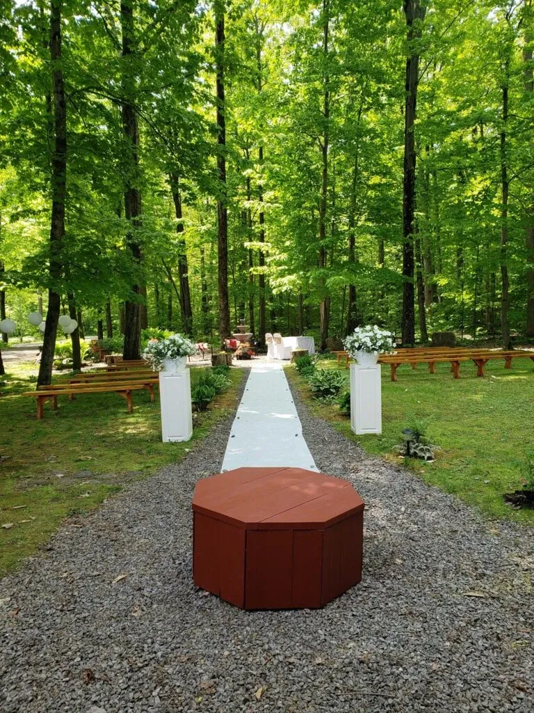 The image showcases an enchanting outdoor wedding setup at L’Érablière D’autrefois Hall, a premier wedding venue in Canada. The aisle is elegantly lined with a white carpet, leading to a beautifully adorned altar surrounded by lush green trees, providing a serene and natural backdrop. Wooden benches are arranged neatly on the grassy area for guests, and floral arrangements with white blossoms enhance the romantic ambiance. The scene is ideal for couples looking to celebrate their special day in a picturesque woodland setting.