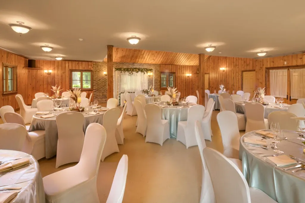 The image shows the interior of L’Érablière D’autrefois Hall, a wedding venue listed on Mariage Elegant in Canada. The spacious room features wooden walls and a high ceiling, creating a warm and inviting atmosphere. Round tables are elegantly set up with white tablecloths and chairs covered in white fabric, adding to the sophisticated decor. Each table is adorned with simple yet tasteful floral centerpieces. Soft lighting enhances the cozy ambiance, making this an ideal setting for a wedding reception.