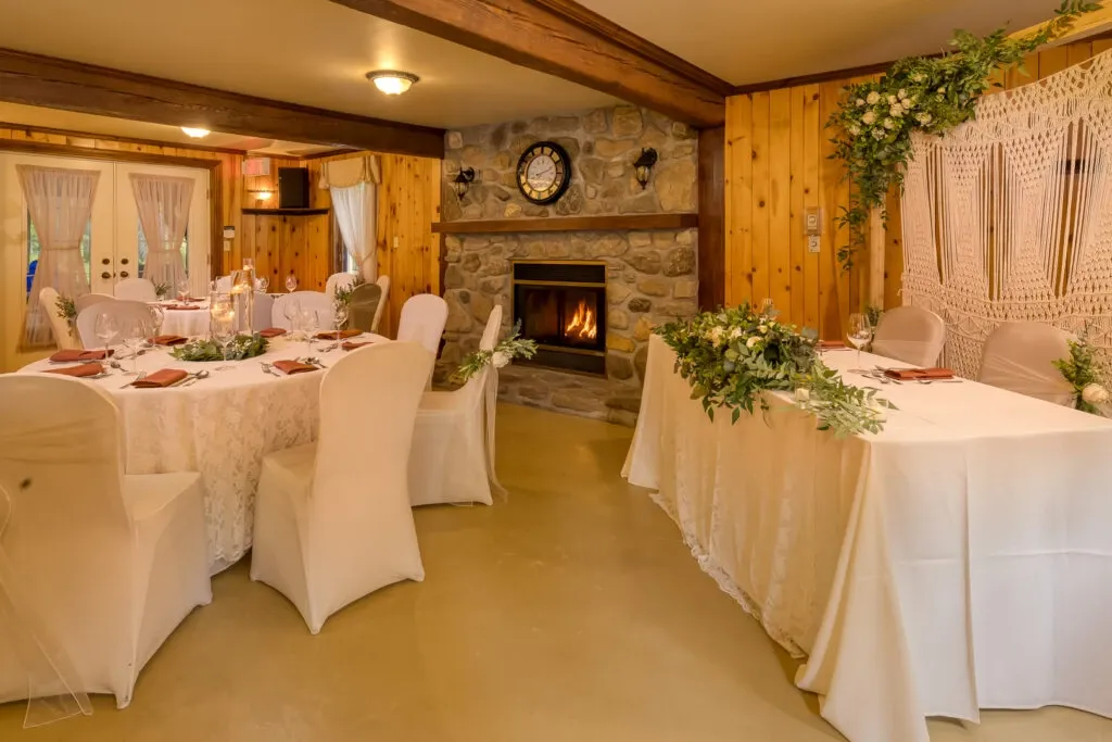 The image features the interior of the L’Érablière D’autrefois Hall, a wedding venue in Canada. The cozy setting includes elegantly decorated tables draped with white cloths and adorned with lush, green floral arrangements. The chairs are covered in soft white fabric, enhancing the venue's sophistication. A warm, stone fireplace is at the center, contributing to the intimate atmosphere of the space. Delicate curtains cover the windows, allowing soft light to filter in, while the walls feature wood paneling, adding a rustic touch. The setting exudes warmth and charm, ideal for a wedding reception.