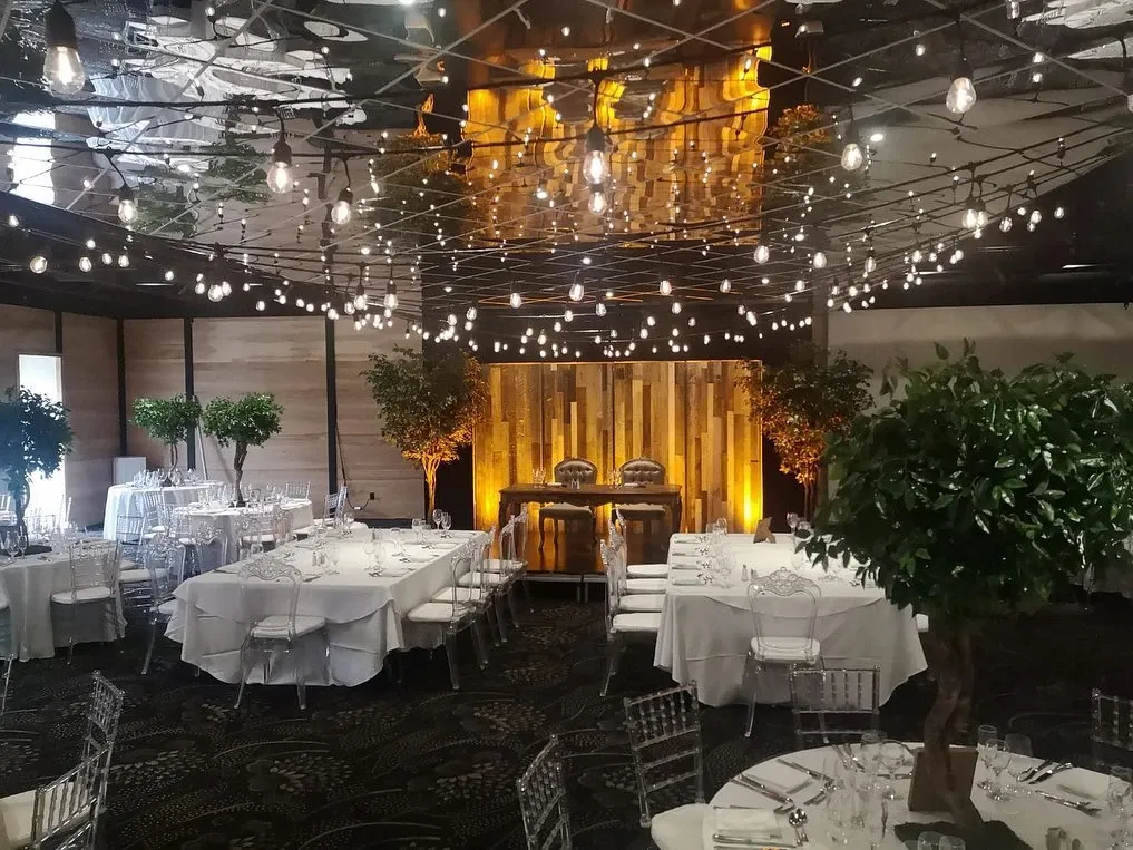 The image showcases an elegantly decorated wedding venue at Plaza Antique, categorized under Wedding Venues. The venue features beautifully arranged tables draped in white linens, each set with fine glassware and silverware, ready to host a sophisticated event. Two lush, green potted trees add a touch of natural elegance to the interior. Overhead, an array of ambient string lights crisscross the ceiling, casting a warm and inviting glow over the room. At the front of the venue, a rustic wooden backdrop highlighted by soft, ambient lighting frames a table set for the wedding couple, emphasizing the romantic atmosphere that Plaza Antique offers for weddings.