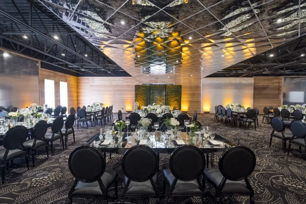 The image shows an elegant wedding venue set up at Plaza Antique, featuring round tables with black cushioned chairs arranged across a spacious, carpeted room. The tables are adorned with white tablecloths, floral centerpieces, and neatly arranged tableware, creating a sophisticated ambiance. Overhead, a striking reflective ceiling adds modern flair, while subtle, warm lighting enhances the inviting atmosphere. This image showcases Plaza Antique as an ideal setting for weddings, highlighting its stylish decor and ample space for hosting memorable events.