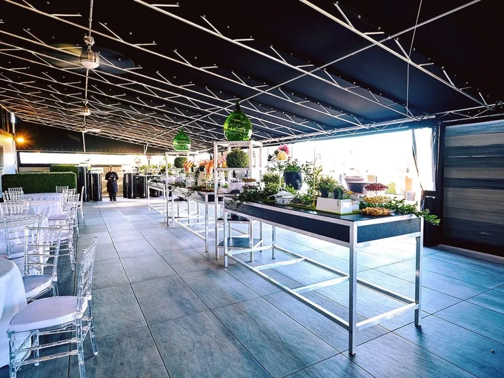 The image shows an elegantly decorated wedding venue at Plaza Antique, categorized under Wedding Venues for Mariage Elegant in Canada. The spacious interior features a contemporary design with a sleek, dark ceiling adorned with crisscrossing lines, adding a modern touch. The venue is set up with clear Chiavari chairs and neatly arranged tables covered in white tablecloths. A series of rectangular tables are artfully arranged with chic centerpieces and greenery, complementing the sophisticated atmosphere. Large windows on the side allow natural light to flood the space, creating an inviting and airy ambiance, ideal for a wedding celebration.