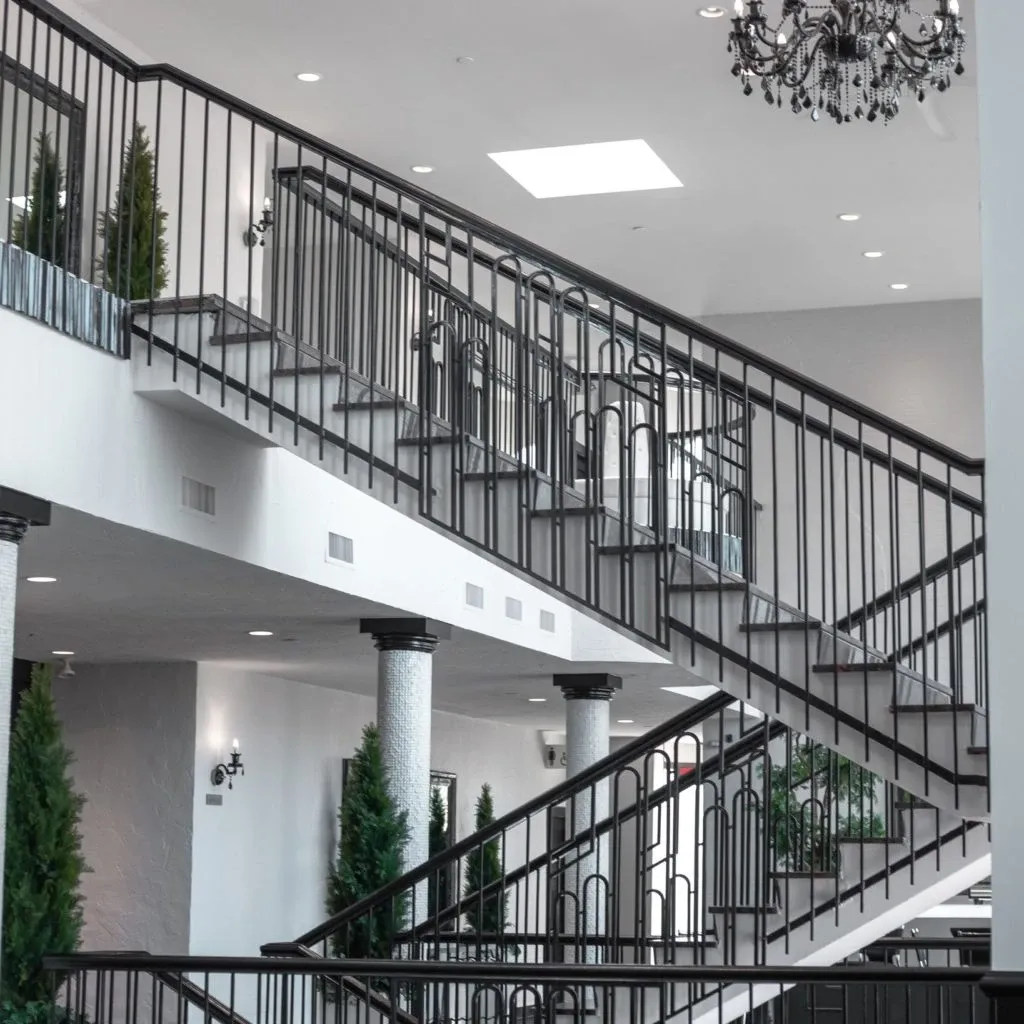 The image showcases an elegant, spacious interior of Plaza Antique, highlighting its suitability as a wedding venue. The photograph features a grand staircase with modern black railings and white steps, flanked by ornate, classical columns. Decorative lush green plants are strategically positioned along the staircase and around the venue, adding a touch of natural beauty. The setting is well-lit, with sophisticated chandeliers providing ambient lighting from the ceiling. This venue offers a stylish and contemporary atmosphere, ideal for hosting weddings in Canada.