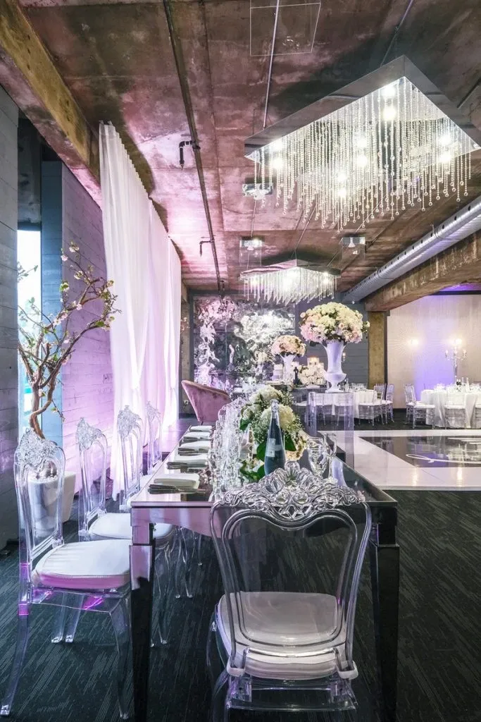 The image showcases a beautifully decorated wedding venue at Plaza Antique, categorized under wedding venues. The setting features elegant, transparent chairs surrounding a sleek, reflective dining table adorned with exquisite floral arrangements. Above, a striking, modern chandelier with cascading lights adds a touch of sophistication. The venue's interior combines industrial elements with lavish décor, including lush, blooming flower centerpieces and ambient mood lighting, creating a romantic and luxurious atmosphere ideal for weddings.