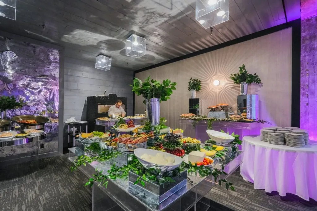 This image shows an elegantly arranged wedding reception setup at Plaza Antique, categorized under Wedding Venues on Mariage Elegant in Canada. The venue features an array of carefully arranged buffet tables adorned with a variety of culinary offerings. Fresh fruits and assorted appetizers are beautifully displayed in white bowls on clear, elevated stands, adding a sophisticated touch. The ambiance is enhanced by subtle purple lighting and decorative greenery, creating a vibrant yet classy atmosphere. A table with stacked white plates is ready for guests, suggesting a well-prepared event setting. The surrounding decor, including modern ceiling lights and textural wall accents, contributes to the chic and inviting environment for wedding celebrations.