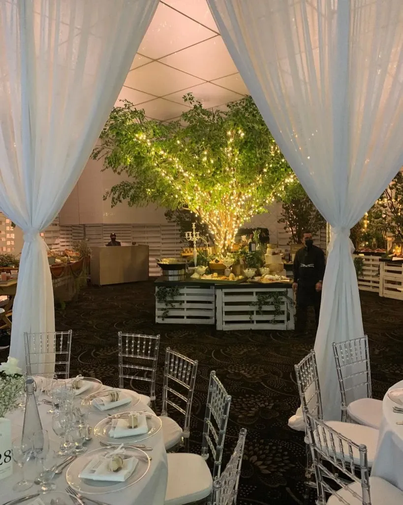 The image showcases a beautifully decorated wedding venue at Plaza Antique, categorized under wedding venues, part of the Mariage Elegant directory in Canada. A large, elegantly lit tree stands at the center, draped with delicate fairy lights, creating a warm and romantic ambiance. Surrounding the tree are white fabric-draped tables set for a wedding, featuring clear glass chargers, neatly folded napkins with placeholders, and floral centerpieces. Clear Chiavari chairs complement the sophisticated setup. White drapes frame the scene, adding to the venue's charm and intimacy, ideal for a memorable wedding celebration.