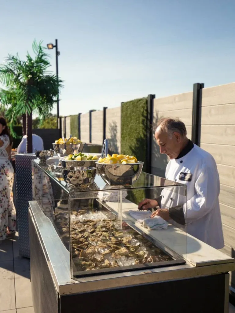 A chef at Plaza Antique, a wedding venue in Canada, is preparing a seafood display on an outdoor patio. The setup includes a variety of fresh oysters arranged on a bed of ice, with bowls of lemons for garnish. In the background, elegantly dressed guests can be seen mingling near a small decorative palm plant, while a blue sky and modern wooden fencing complete the serene ambiance. This vibrant scene highlights the catering options available for weddings at Plaza Antique.