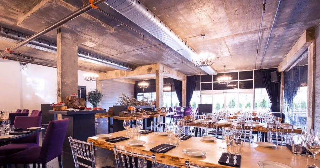 The image showcases Plaza Antique, a featured wedding venue on Mariage Elegant, Canada. The venue's interior is elegantly set up for a wedding reception, featuring a spacious room with industrial-style ceilings and exposed pipes. The tables are adorned with clear, modern glassware and neatly folded black napkins. Purple-cushioned chairs and transparent acrylic chairs offer diverse seating options. The ambiance is enhanced by soft lighting from sophisticated chandeliers, casting a warm glow over the room. Large windows draped with dark curtains allow natural light to fill the space, adding a touch of elegance to the industrial decor.