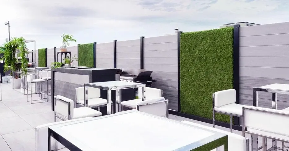 The image showcases an elegant outdoor setting at Plaza Antique, a renowned wedding venue listed under wedding vendors on Mariage Elegant, Canada. The scene features modern white tables and chairs arranged on a sleek, spacious patio. The area is bordered by contemporary grey and green privacy panels, providing a chic and intimate ambiance perfect for wedding receptions or ceremonies. Lush greenery accents the space, enhancing the fresh and inviting atmosphere ideal for celebrating special occasions.