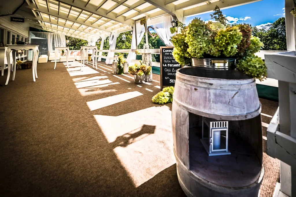 The image showcases an elegant wedding venue by La Distinction, categorized under Wedding Venues on Mariage Elegant, Canada. It features an inviting outdoor terrace with a rustic charm, highlighted by a large wooden barrel, which adds a touch of vintage decor. The space is beautifully adorned with lush arrangements of green and cream flowers, creating a romantic and natural ambiance. Bright sunlight filters through a partially covered roof, casting artistic shadows on the floor and illuminating the seating area, which is set up with white tables and chairs, ideal for a picturesque wedding reception.