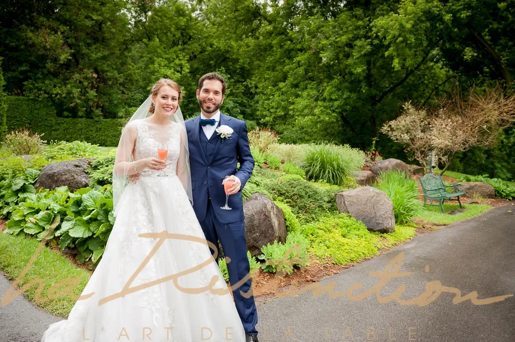 A bride and groom are standing together in a lush garden setting at the wedding venue La Distinction, showcasing the romantic ambiance of this Canadian location. The bride is wearing an elegant white lace gown and veil, while the groom is dressed in a formal navy suit with a boutonniere. Both are smiling and holding glasses with a light pink beverage, possibly for a toast. Behind them are vibrant green plants and stones, creating a picturesque backdrop suitable for wedding photography. This image highlights the enchanting outdoor setting available at La Distinction for weddings.